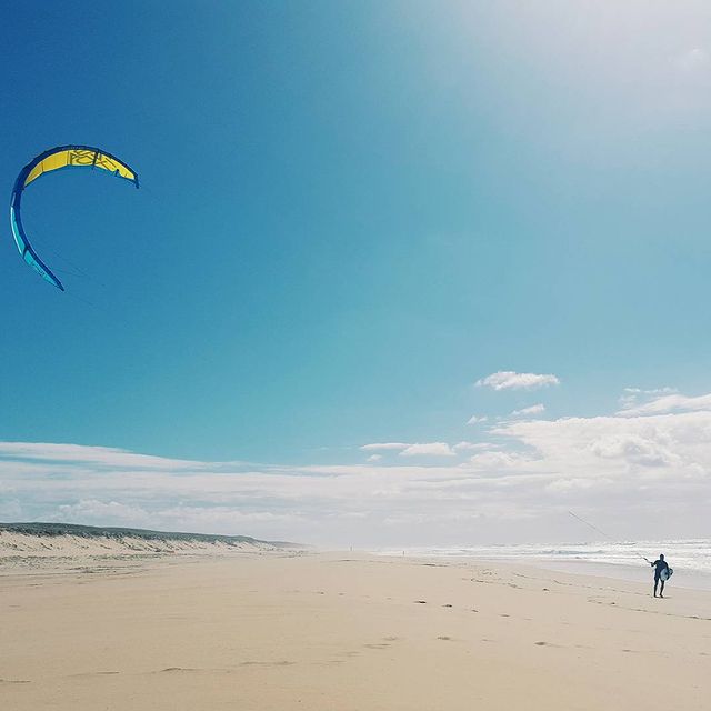 plage au Cap Ferret, ciel bleu, un homme porte un kite-surf dans un bras, une voile de kite surf dans l'autre