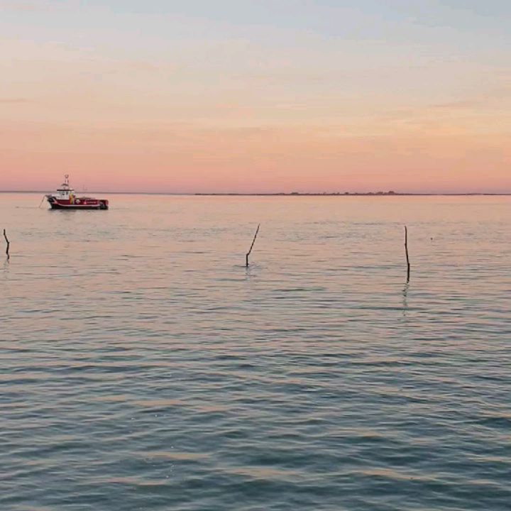 bassin d'Arcachon au coucher du soleil, ciel orangé, un bateau de pêche à coque rouge rentre au port