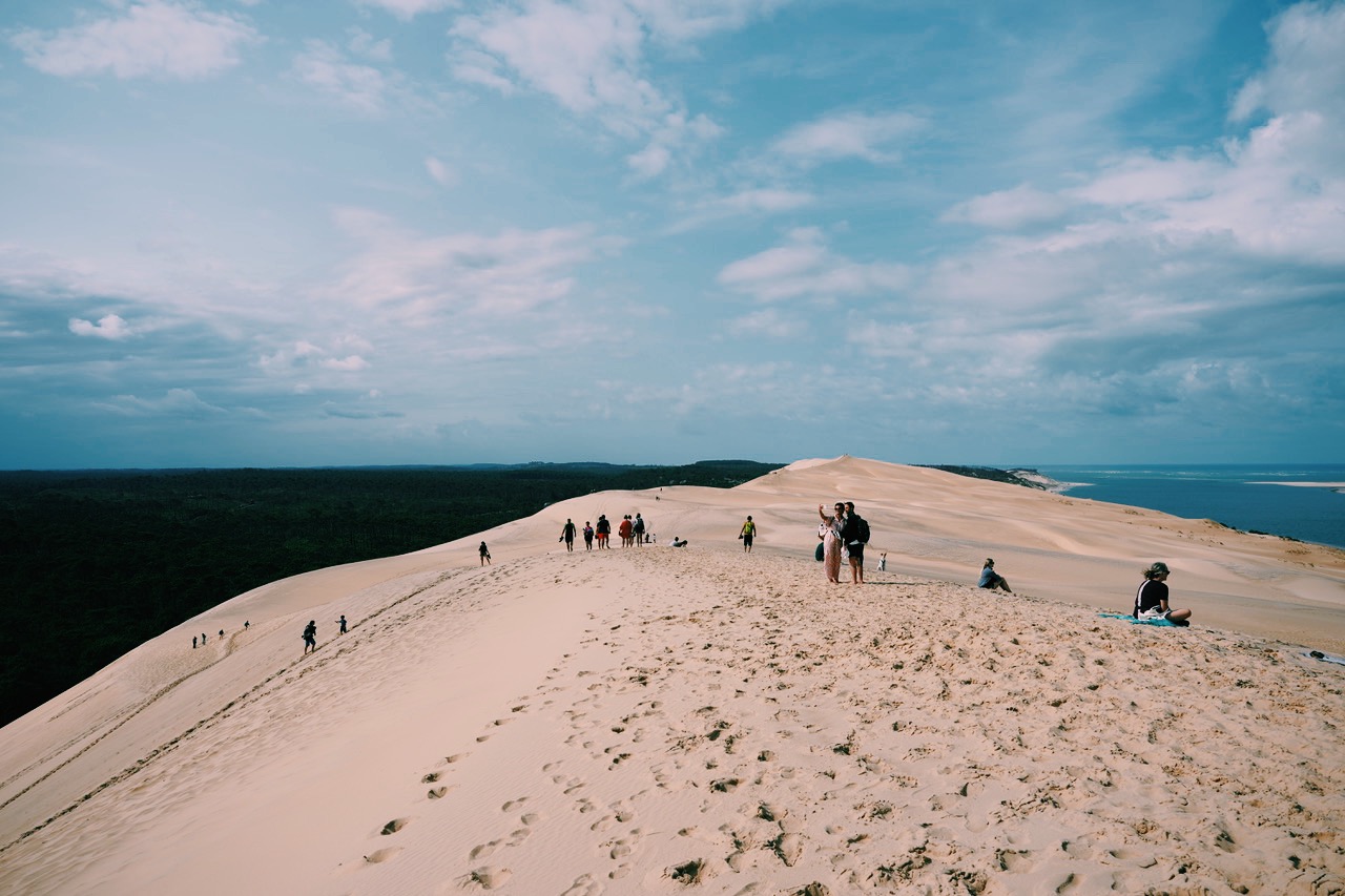 Photo prise sur la Grande Dune du Pilat, à gauche la forêt, à droit l'océan, ciel bleu avec nuages, quelques personnes sur la Dune dont un couple au milieu qui se prend en selfie